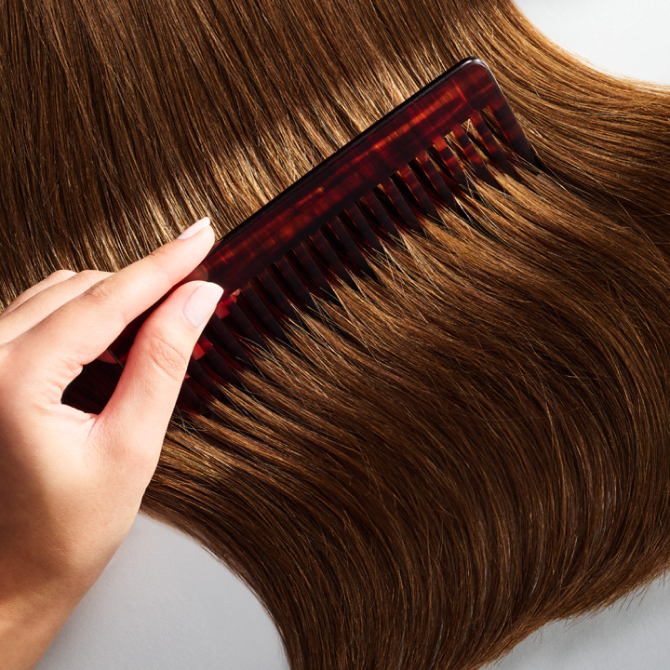 A woman with long brown hair gently combs her hair with a comb.